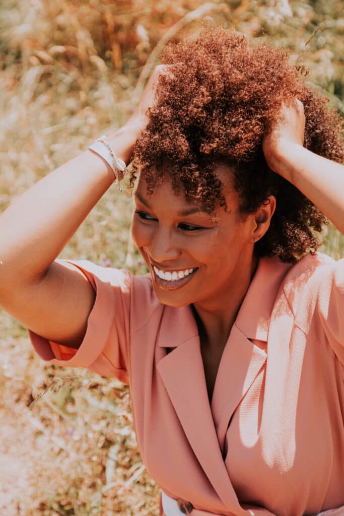 Une femme noire passe la main dans ses cheveux crépus, elle est souriante et se tient dans un champ de blé.