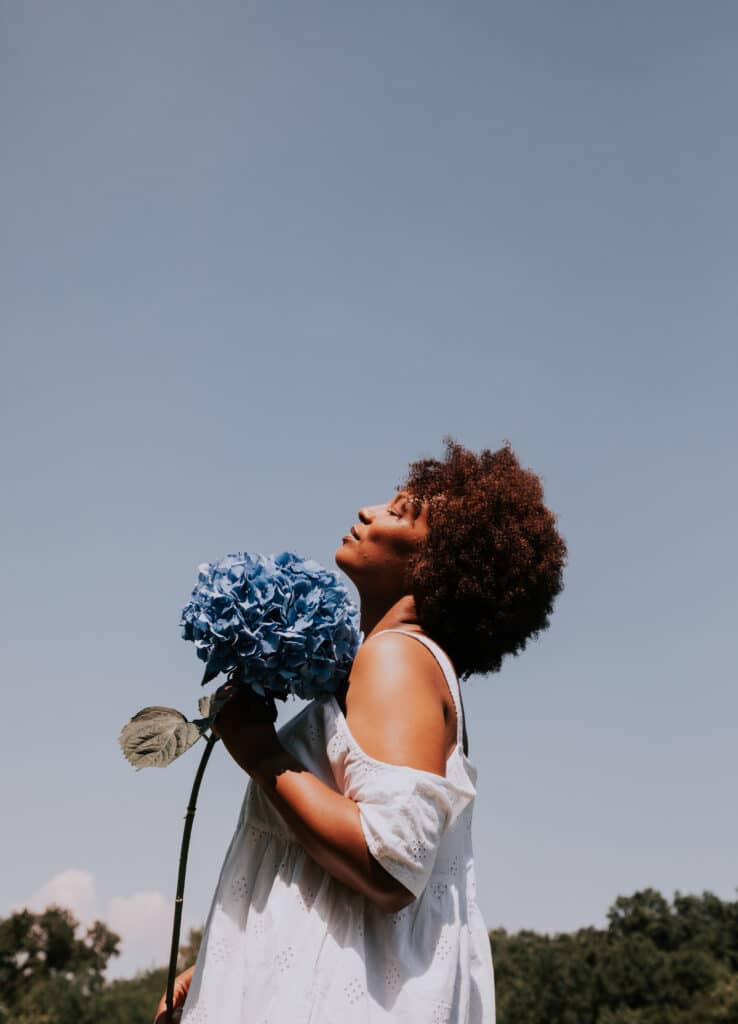 Une femme noire se tient de profil, la tête légèrement levée vers le ciel, ses cheveux crépus en arrière. Elle tient une fleur bleu entre ses mains et semble sereine.