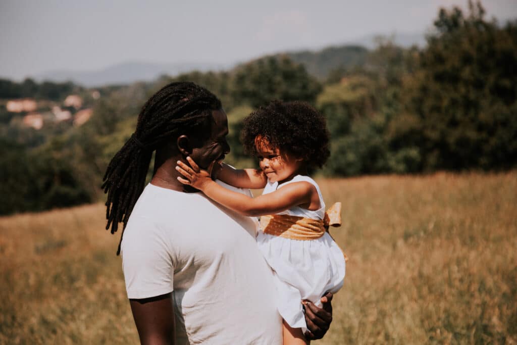 Un papa tient sa fille entre ses bras, ils se regardent avec complicité en souriant. Le père a ses cheveux attachés et sa fille a les cheveux crépus détachés